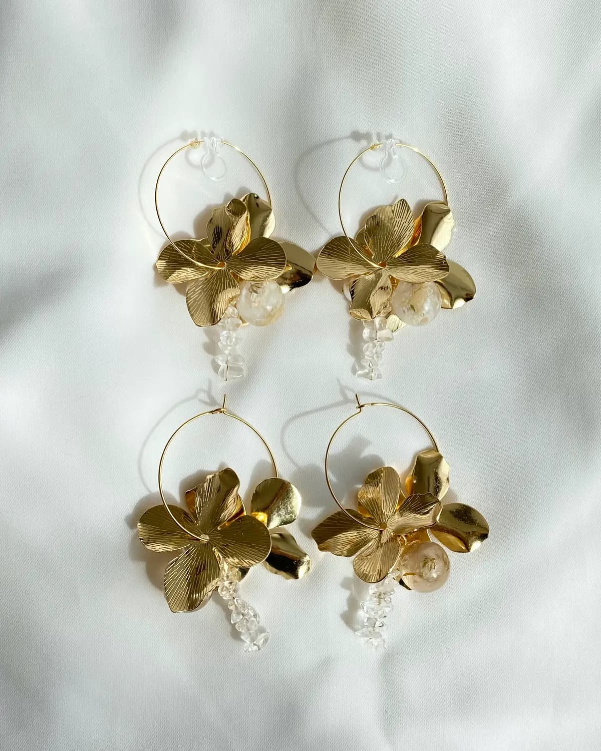 白いサテンの上に並ぶ、金の花びらのフープピアス2組。花芯にはドライフラワーをとじこめた透明な球と、透明ビーズの飾りが揺れる