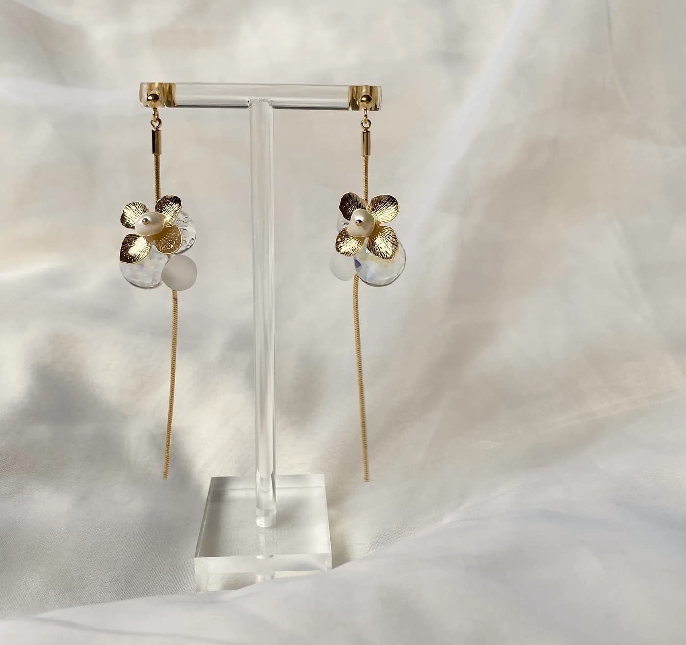 白い布を背景に、アクリルスタンドで揺れるikyuのピアス。金の花びらから、白い小花と細いゴールドのラインが垂れる